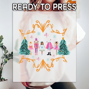 May include: A white t-shirt with a watercolor design of the Nutcracker and his friends. The design features a gold ornate frame with a green Christmas tree on each side. The characters are dressed in colorful costumes and are holding a white mouse.