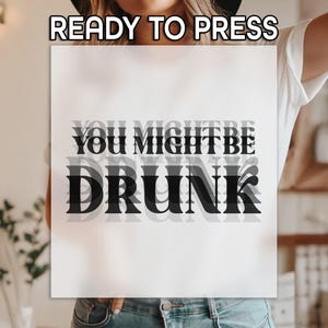 May include: A white sign with the text "READY TO PRESS" at the top. Below, the words "YOU MIGHT BE DRUNK" are layered in a black, stylized font, creating a blurred effect. The sign is held by a person.