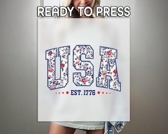 May include: White t-shirt with a blue and red floral print of the word "USA" and the text "EST. 1776" below.
