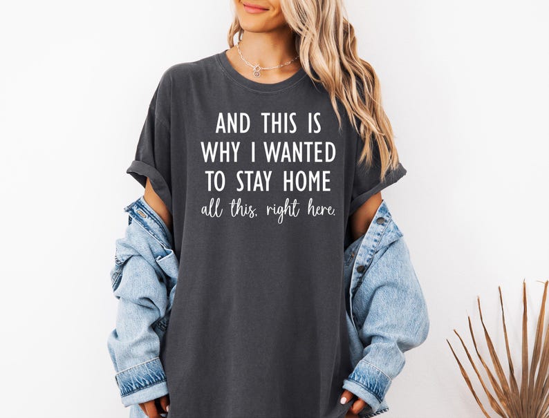 May include: Dark gray t-shirt with the text "AND THIS IS WHY I WANTED TO STAY HOME all this, right here." The shirt has rolled-up sleeves and a relaxed fit. A denim jacket is worn over the shirt.