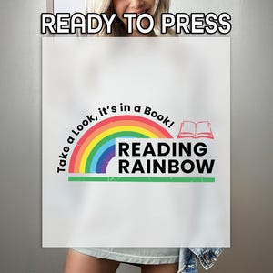 May include: White sublimation transfer with the text "READY TO PRESS" at the top. The design features a rainbow arch with the words "Take a Look, it's in a Book!" and "READING RAINBOW" in black. A red open book graphic is also included.
