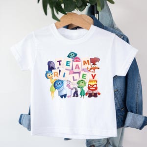 May include: White t-shirt featuring the phrase "TEAM RILEY" in colorful block letters, surrounded by cartoon characters. The shirt is hanging on a wooden hanger against a denim jacket.