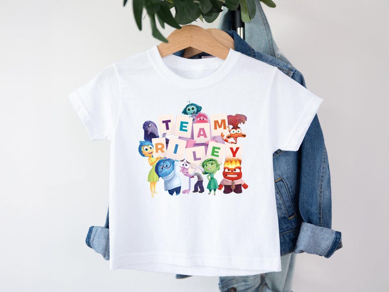 May include: White t-shirt featuring the phrase "TEAM RILEY" in colorful block letters, surrounded by cartoon characters. The shirt is hanging on a wooden hanger against a denim jacket.