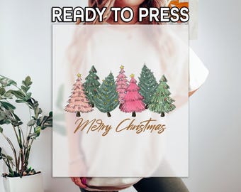 May include: A white t-shirt with a design of six stylized Christmas trees in shades of green, pink, and white. The trees are decorated with gold stars and the text "Merry Christmas" is printed in gold below the trees.