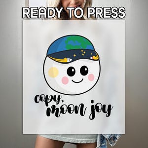 May include: A white circular graphic featuring a smiling face with pink cheeks and a yellow spot. The character wears a blue and black cap with a planet design and yellow stars. Below the character, the black text reads, copy, moon joy.