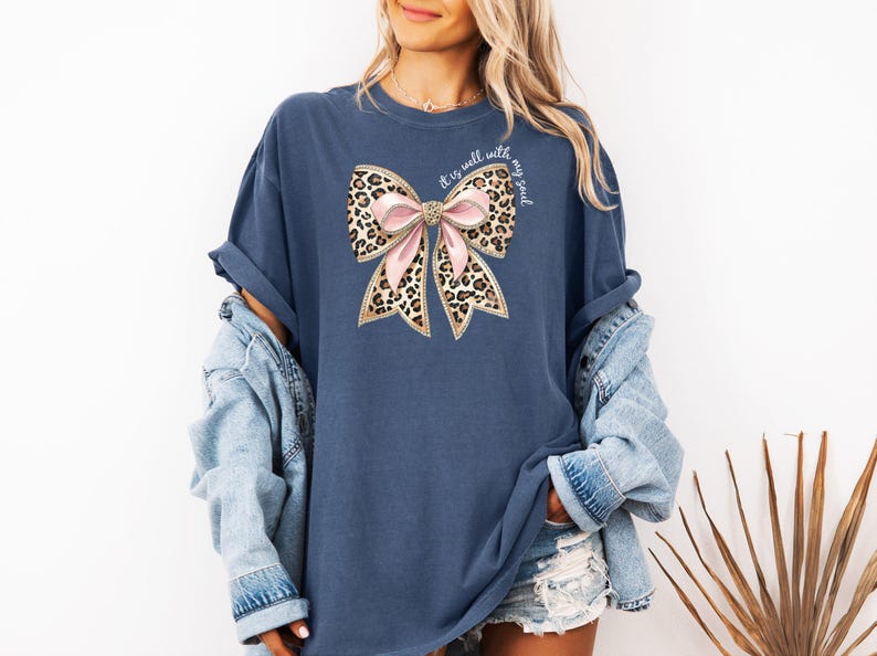 May include: A navy blue t-shirt with a large pink and leopard print bow with gold glitter accents. The text "It is well with my soul" is written in white on the bow.