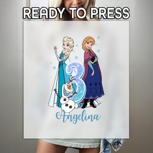 May include: A white sublimation transfer featuring Elsa and Anna from Frozen, with the number 3 in blue glitter and the name "Angelina" in script. The text "READY TO PRESS" is at the top.