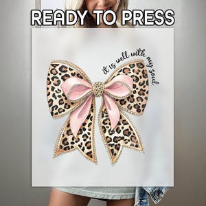 May include: A white t-shirt with a pink and brown leopard print bow with rhinestones and the text "It is well with my soul".