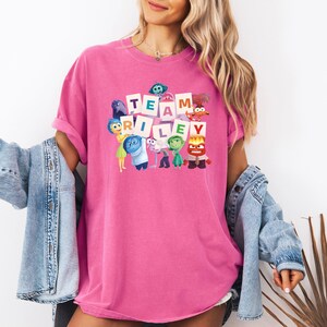 May include: A hot pink t-shirt with the words "TEAM RILEY" in white block letters. The shirt features cartoon characters from the movie Inside Out. The shirt is a comfortable, oversized fit.