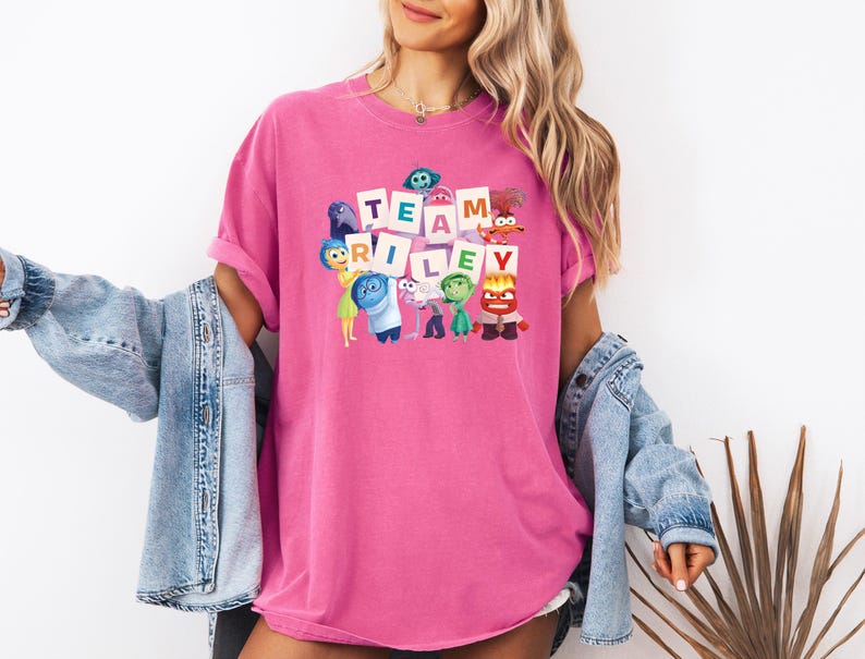 May include: A hot pink t-shirt with the words "TEAM RILEY" in white block letters. The shirt features cartoon characters from the movie Inside Out. The shirt is a comfortable, oversized fit.