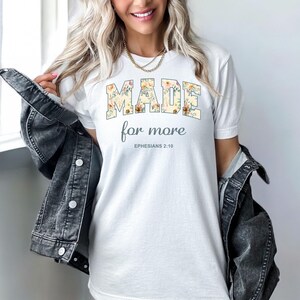 Op de afbeelding: Wit T-shirt met een bloemenontwerp met de tekst "MADE for more" en "EPHESIANS 2:10" in grijze tekst.