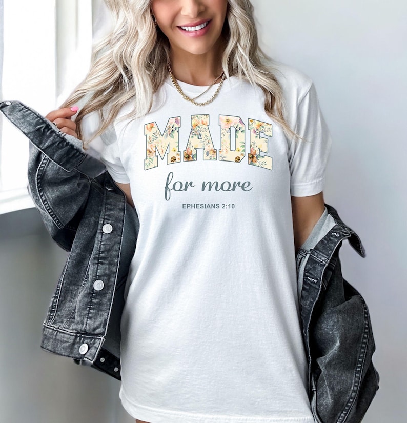 Op de afbeelding: Wit T-shirt met een bloemenontwerp met de tekst "MADE for more" en "EPHESIANS 2:10" in grijze tekst.