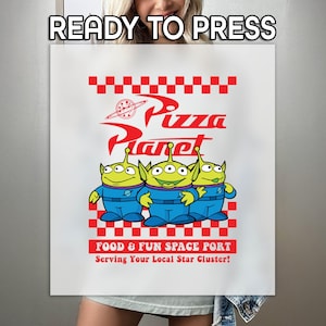 May include: White t-shirt with a red and white checkered background and a graphic of three green aliens from the movie Toy Story. The graphic says "Pizza Planet" in red letters and "Food & Fun Space Port" in red letters. The text below says "Serving Your Local Star Cluster!"