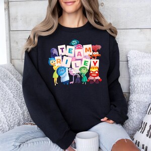 May include: Black crewneck sweatshirt featuring the words "TEAM RILEY" in a block letter design, with cartoon characters from the movie Inside Out. The sweatshirt is worn by a person sitting on a couch with pillows.