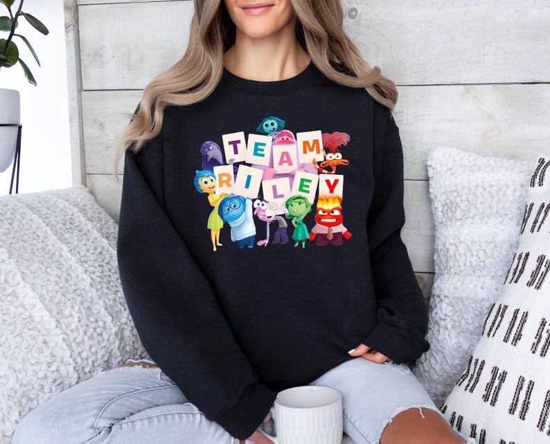 May include: Black crewneck sweatshirt featuring the words "TEAM RILEY" in a block letter design, with cartoon characters from the movie Inside Out. The sweatshirt is worn by a person sitting on a couch with pillows.