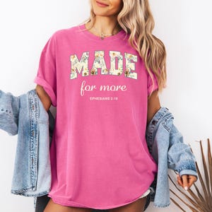Op de afbeelding: Roze T-shirt met een bloemenprint die "MADE for more" en "EPHESIANS 2:10" in witte tekst zegt.