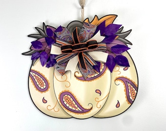 May include: A white pumpkin door hanger with a paisley pattern and purple and orange accents. A large purple and black bow with a paisley pattern is attached to the top of the pumpkin.