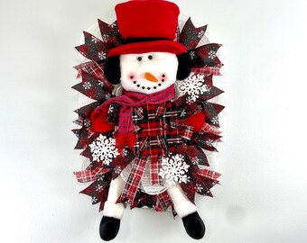 May include: A snowman wreath featuring a red hat, black eyes, and a carrot nose. The wreath is adorned with red and black plaid ribbon, white snowflakes, and white mesh.