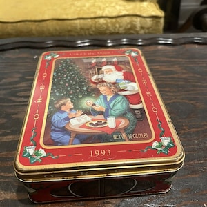 Op de afbeelding: Rode en gouden metalen doos met een kerstscène met de kerstman, een kind en een kerstboom. De doos heeft de tekst "Unlocking Magic!" en "1993" erop. De doos heeft een nettogewicht van 16 ounce (1 lb).
