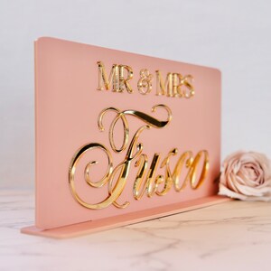 Peut inclure: Panneau de mariage en acrylique rose blush avec lettrage dor&eacute; miroir. Le panneau indique "MR & MRS" au-dessus du nom de famille "Fusco" en &eacute;criture &eacute;l&eacute;gante. Une seule rose repose &agrave; c&ocirc;t&eacute; du panneau.