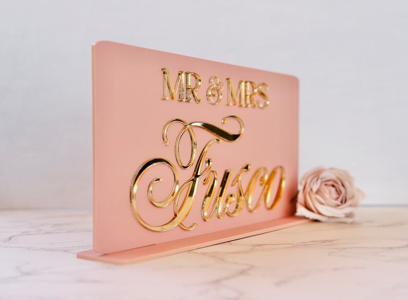 Peut inclure: Panneau de mariage en acrylique rose blush avec lettrage dor&eacute; miroir. Le panneau indique "MR & MRS" au-dessus du nom de famille "Fusco" en &eacute;criture &eacute;l&eacute;gante. Une seule rose repose &agrave; c&ocirc;t&eacute; du panneau.