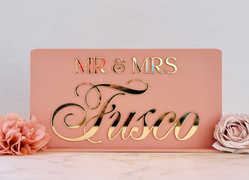 Peut inclure: Panneau de mariage rose blush avec lettrage dor&eacute; miroir. Le panneau indique "MR & MRS" au-dessus du nom de famille "Fusco" dans une &eacute;criture &eacute;l&eacute;gante. Des fleurs d&eacute;coratives sont au premier plan.