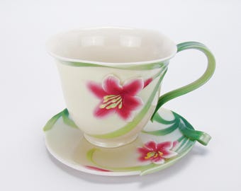 Puede incluir: Un juego de taza y platillo de té de porcelana blanca con un diseño floral. La taza y el platillo presentan una flor rosa con hojas verdes.