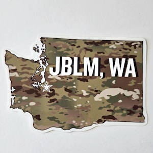 May include: A sticker in the shape of the state of Washington with a camouflage pattern. The text "JBLM, WA" is printed in bold white letters across the center of the sticker. A white star is located near the bottom of the text.
