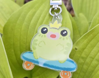 May include: A light green frog wearing a crown, riding a blue skateboard with orange wheels. The frog keychain has a silver clasp and is set against a backdrop of green leaves.