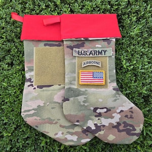 May include: A pair of camouflage Christmas stockings with red cuffs. The stockings have patches that say "U.S. ARMY" and "AIRBORNE" with an American flag patch.