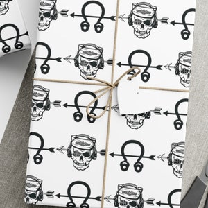 Può includere: Carta da regalo bianca con un designo di teschio e freccia in bianco e nero. Il teschio indossa le cuffie e la freccia punta a destra. La carta è avvolta attorno a una scatola regalo.