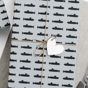 Puede incluir: Tres hojas de papel de regalo con un patrón repetitivo de submarinos negros sobre un fondo gris claro. El papel de regalo está atado con cuerda y decorado con estrellas y corazones de papel blanco.