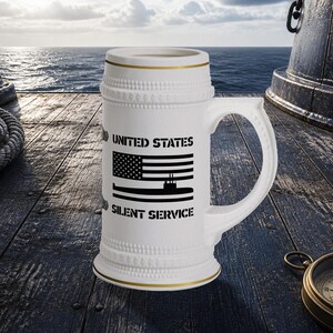 May include: A white ceramic beer stein featuring a black graphic of the United States flag above a submarine silhouette, with the text UNITED STATES SILENT SERVICE printed in black. The stein has a textured rim and base with gold trim accents.