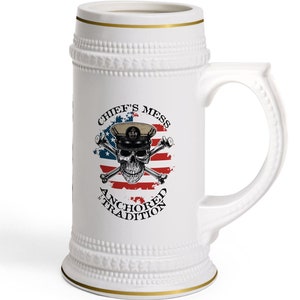 May include: White ceramic beer stein with a gold rim and a design featuring a skull wearing a navy hat with crossed bones and an American flag. The text "Chief's Mess Anchored Tradition" is printed below the skull.