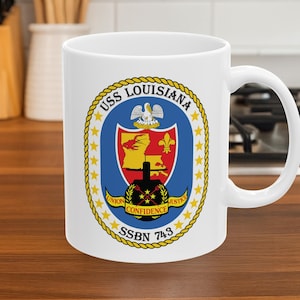 May include: A white ceramic mug featuring the USS Louisiana SSBN 743 emblem. The circular logo includes a yellow map of Louisiana, a red shield, a pelican, a black submarine silhouette, and the text Union, Confidence, Justice in a gold banner.