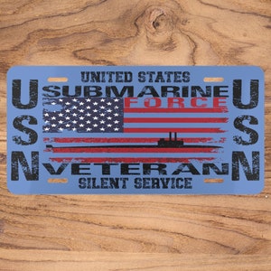 Puede incluir: Una placa de metal azul con el texto "United States Submarine Force USN Veteran Silent Service" y una bandera estadounidense desgastada con la silueta de un submarino.
