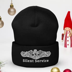 Puede incluir: Un gorro negro con un diseño bordado en blanco y el texto "Silent Service". El gorro se muestra con adornos navideños, incluyendo bolas y pequeñas figuras.