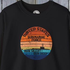 Puede incluir: Sudadera negra con un diseño gráfico circular. El diseño presenta el texto "United States Submarine Force" en blanco y la frase "Run Silent Run Deep" en azul. El diseño tiene rayas naranjas, amarillas y azules, que recuerdan a una puesta de sol.
