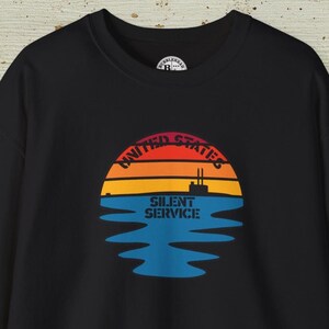 Puede incluir: Sudadera negra con un gráfico de una puesta de sol sobre el agua. El diseño incluye las palabras "UNITED STATES" y "SILENT SERVICE". La puesta de sol es en rojo, naranja y amarillo, con una silueta de barco en el centro.