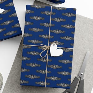 Puede incluir: Una caja de regalo envuelta con papel de regalo azul marino con un patrón repetido de insignias de submarinos plateadas y grises. La caja está atada con hilo y una etiqueta blanca en forma de corazón. También se ven tijeras y otros regalos envueltos.