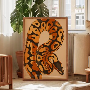 Peut inclure: Une affiche encadrée représentant une illustration stylisée d'un serpent. Le serpent est représenté en nuances d'orange, de marron et de noir sur un fond beige. L'œuvre est exposée dans une pièce avec de la lumière naturelle et une grande plante.