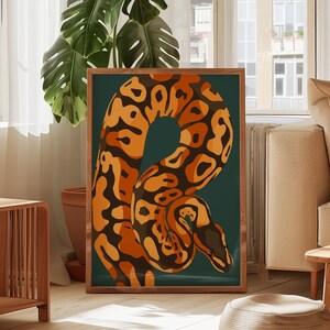 Peut inclure: Une affiche encadrée représentant une illustration stylisée d'un serpent. Le serpent est représenté dans des tons orange et marron sur un fond vert foncé. L'œuvre est exposée dans une pièce avec de la lumière naturelle et une grande plante d'intérieur.