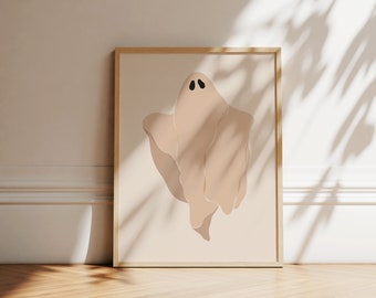 May include: A framed wall art print featuring a stylised, beige-coloured ghost illustration with two simple black oval eyes. The artwork is set against a solid light beige background and is enclosed within a thin, light-toned wooden frame.