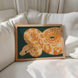 May include: A framed art print featuring a stylized illustration of a snake. The snake is depicted in shades of orange and cream against a dark teal background. The wooden frame adds a natural touch to the artwork, which is displayed on a white surface.