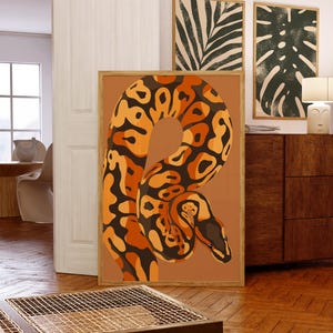 Puede incluir: Una lámina de arte mural rectangular enmarcada que presenta una ilustración estilizada de una serpiente con un cuerpo estampado en tonos naranja, marrón y negro. La serpiente está enroscada sobre un fondo sólido de color terracota en un marco de madera.