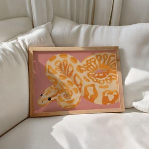 Peut inclure: Une impression d'art encadrée représentant une illustration stylisée d'un serpent. Le serpent est représenté dans des tons orange et crème sur un fond rose. L'œuvre est encadrée dans un cadre en bois brun clair, pour la décoration intérieure.