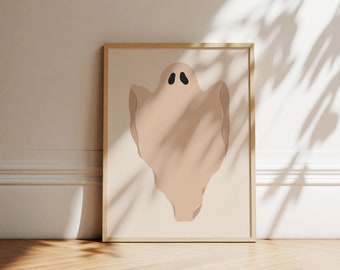 May include: A framed wall art print featuring a beige ghost illustration with two black oval eyes centred on a light tan background. The artwork is enclosed in a simple, light-coloured wooden frame, presenting a clean and straightforward graphic design.