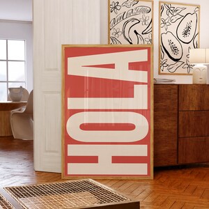 Op de afbeelding: Een grote, ingelijste print met het woord "Aloha" in witte letters op een koraalroze achtergrond. De print hangt aan een witte muur naast twee kleinere ingelijste prints met zwart-wit illustraties van tropisch fruit.