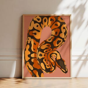 May include: A framed art print showing a stylized snake illustration. The snake is rendered in orange, brown, and black against a pink backdrop. The artwork is displayed in a room with natural light and a wooden floor.