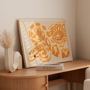 May include: A framed art print featuring an abstract illustration of a snake in shades of orange and tan. The artwork is displayed on a wooden table, with a vase and a book titled "MATISSE" visible.
